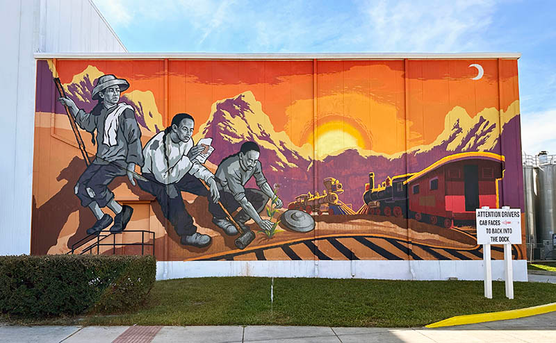 A colorful mural painted on a wall in a city. Three Chinese workers sit on a hillside.