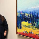 Woman standing beside a colorful landscape painting.