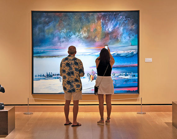 Two people stand in front of a large colorful landscape painting in an art gallery; one person is taking a photo with a smartphone.