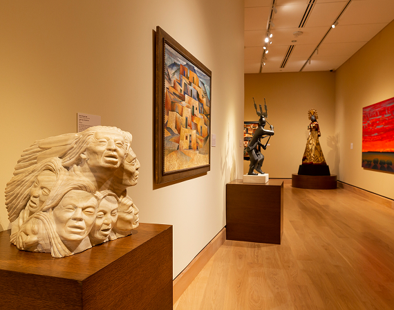 Art gallery interior with sculptures and paintings on beige walls; wooden floor, spotlights, and various art pieces displayed on pedestals and walls.