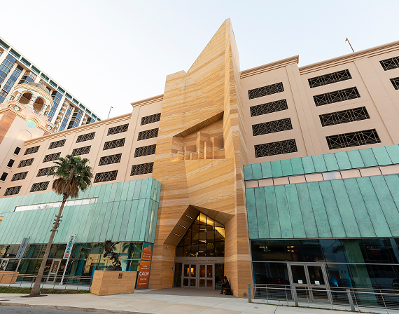 Modern building with tan stone geometric facade, green glass panels at street level, and multiple large windows; palm tree in foreground.