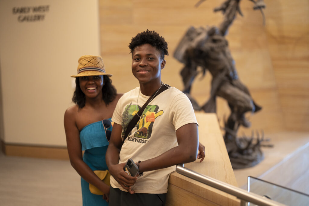 Two people stand indoors near a railing, smiling at the camera. A large abstract sculpture and a sign reading "EARLY WEST GALLERY" are visible in the background.