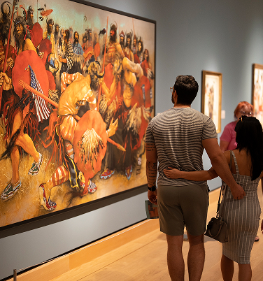 A couple stands arm-in-arm in an art gallery, viewing a large, colorful painting depicting a group of warriors in battle.