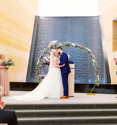 A bride and groom share a kiss under a floral arch during their wedding ceremony, with bridal party members standing nearby.