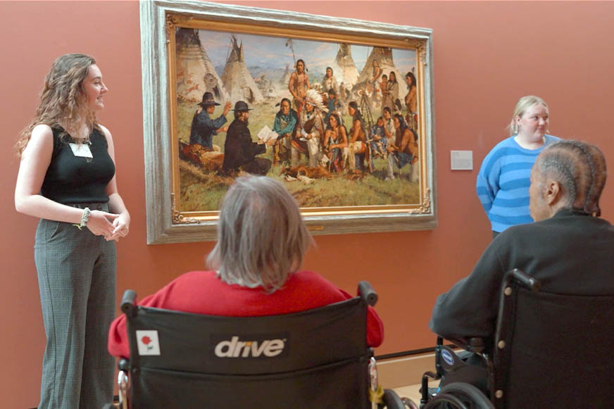 A museum guide speaks to visitors, including two people in wheelchairs, in front of a large painting of a Native American scene, highlighting the museum’s Accessibility Programs.