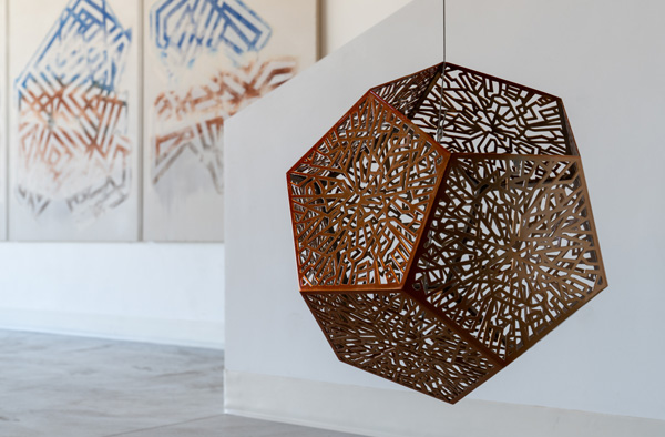 A geometric metal sculpture with intricate cut-out patterns hangs indoors, offering a cross-continental reflection, while abstract artwork from the West is visible on the wall behind it.