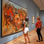 Visitors observing large, vibrant historical painting in gallery.