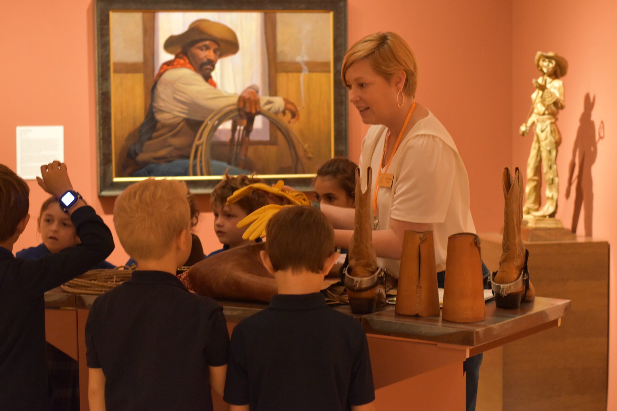 A museum guide leads children on educational field trips, showing cowboy gear like boots and gloves in front of a Western-themed painting and statue.