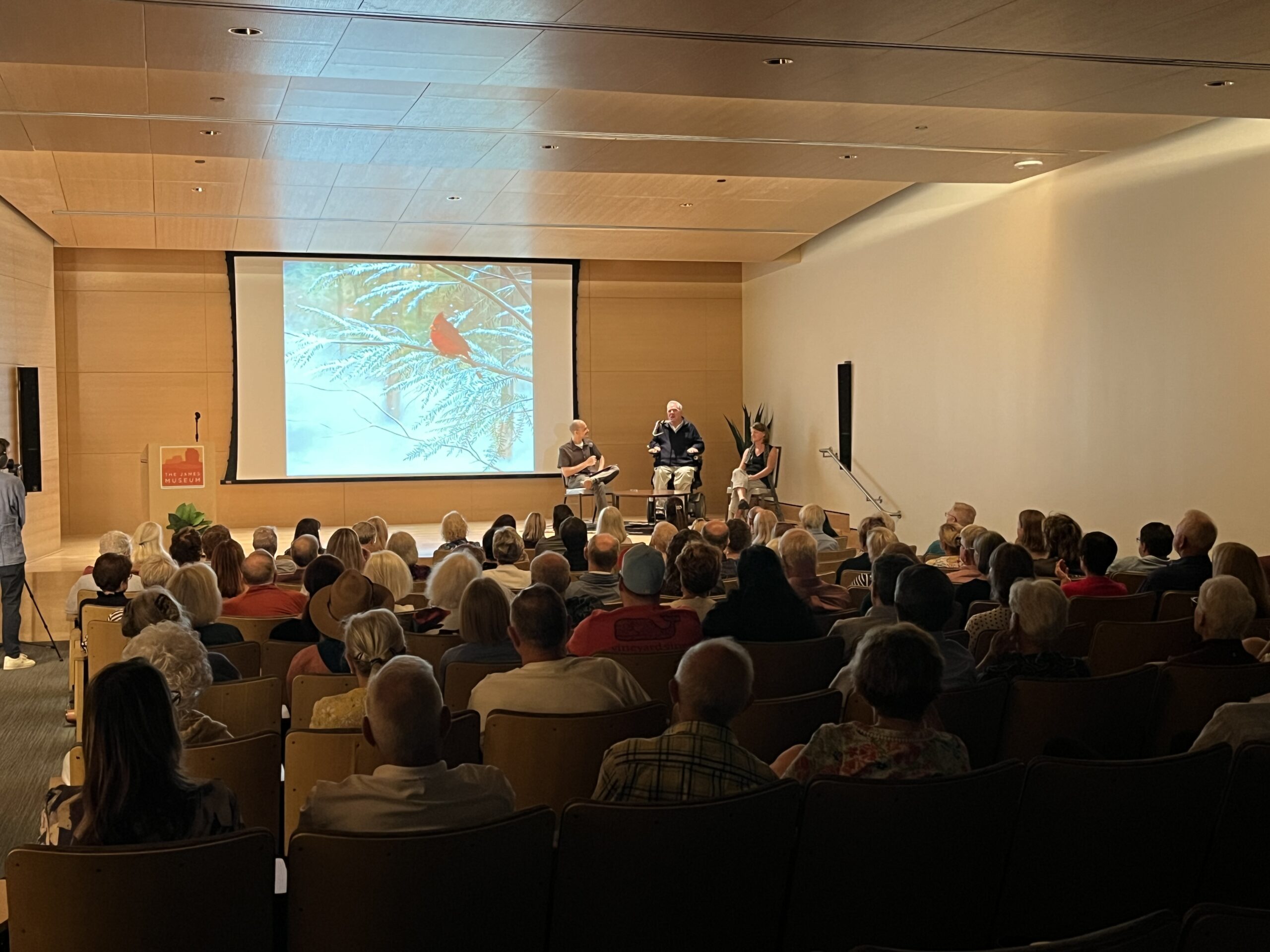 Audience seated in an auditorium Event Space watching a panel discussion; a projected image of a cardinal on a snowy branch is displayed behind the speakers.