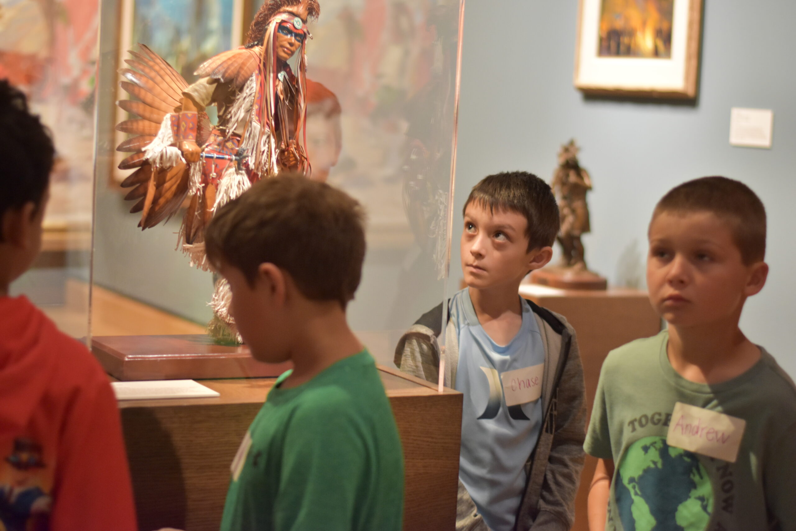 Four boys stand in a museum, looking at a Native American regalia display behind glass during one of their field trips.