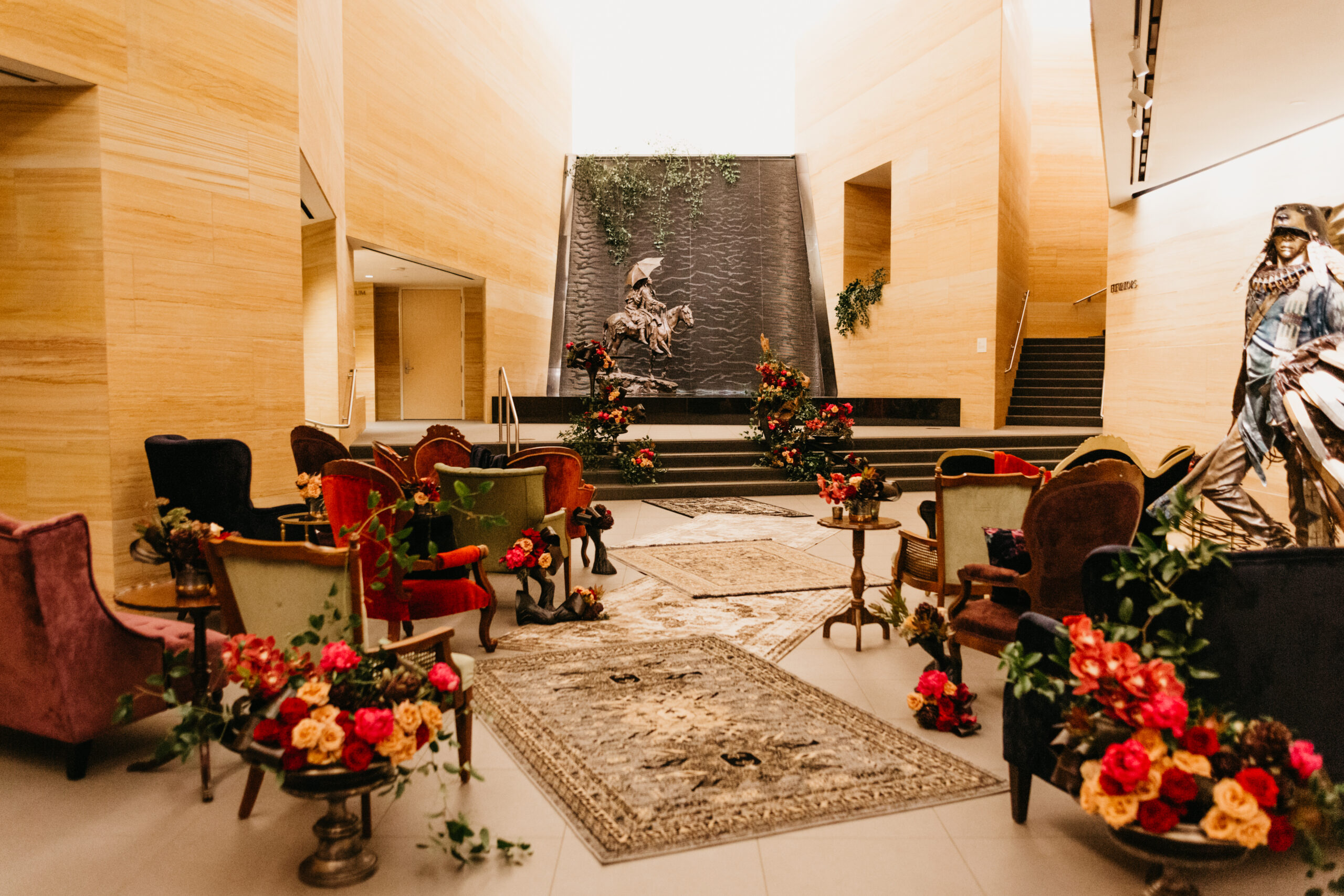 A modern lobby with vintage armchairs, rugs, and floral arrangements serves as an inviting event space, featuring a black wall fountain adorned with a horse statue and lush greenery.