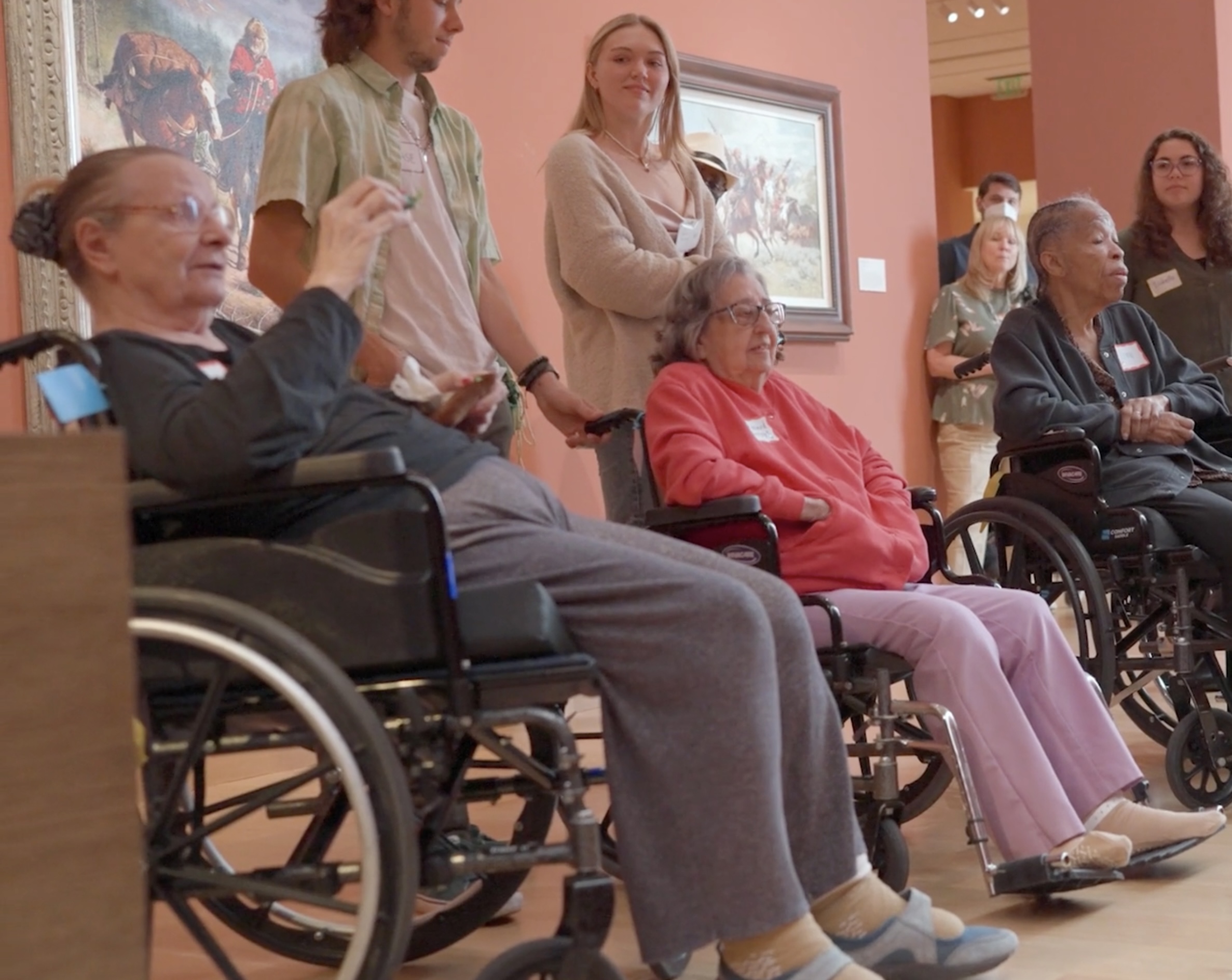 Three elderly women in wheelchairs sit in an art museum, accompanied by younger individuals standing behind them. Paintings grace the walls, highlighting the importance of art appreciation—consider making a donation to support such enriching experiences.