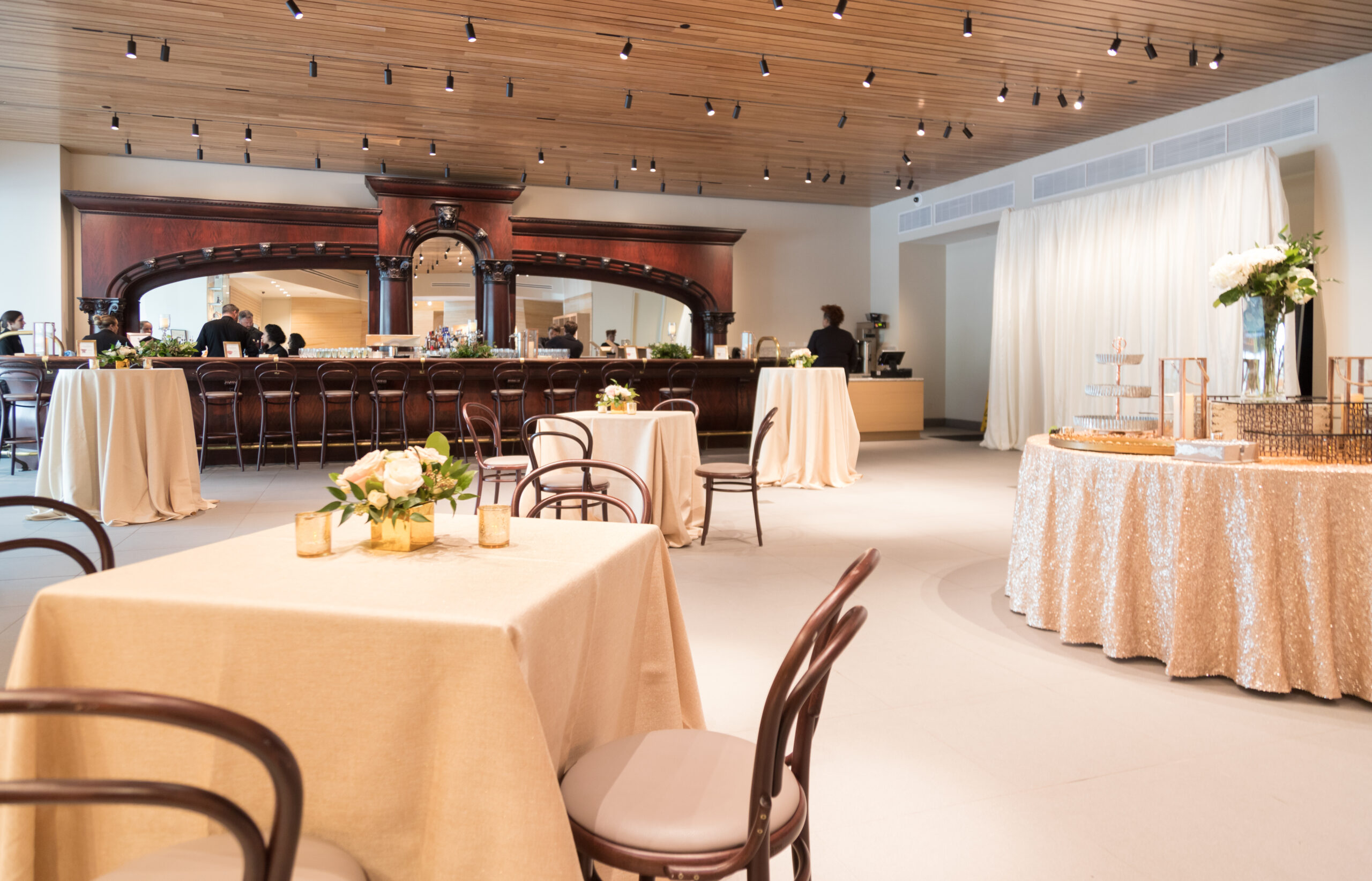 A spacious, elegant event venue—ideal for those seeking premium event spaces—features round and square tables with beige cloths, floral centerpieces, and a large wooden bar in the background.