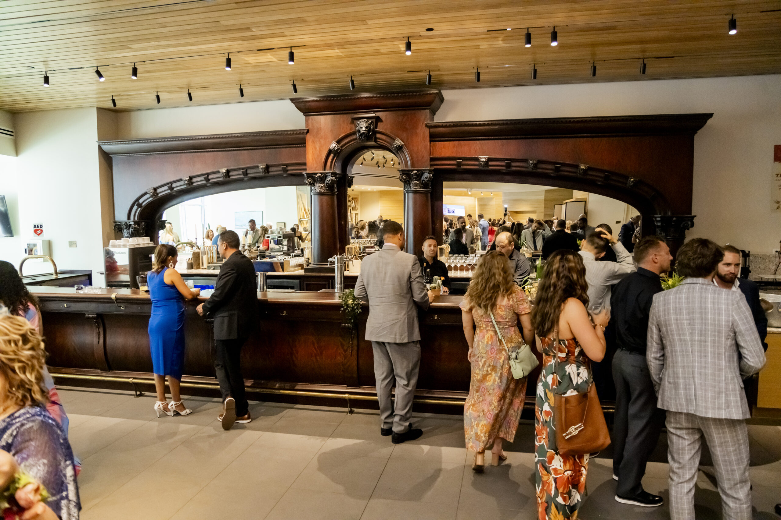 People in formal and semi-formal attire gather and socialize at a large wooden bar inside one of the city’s premier event spaces, enjoying the spacious, well-lit venue.