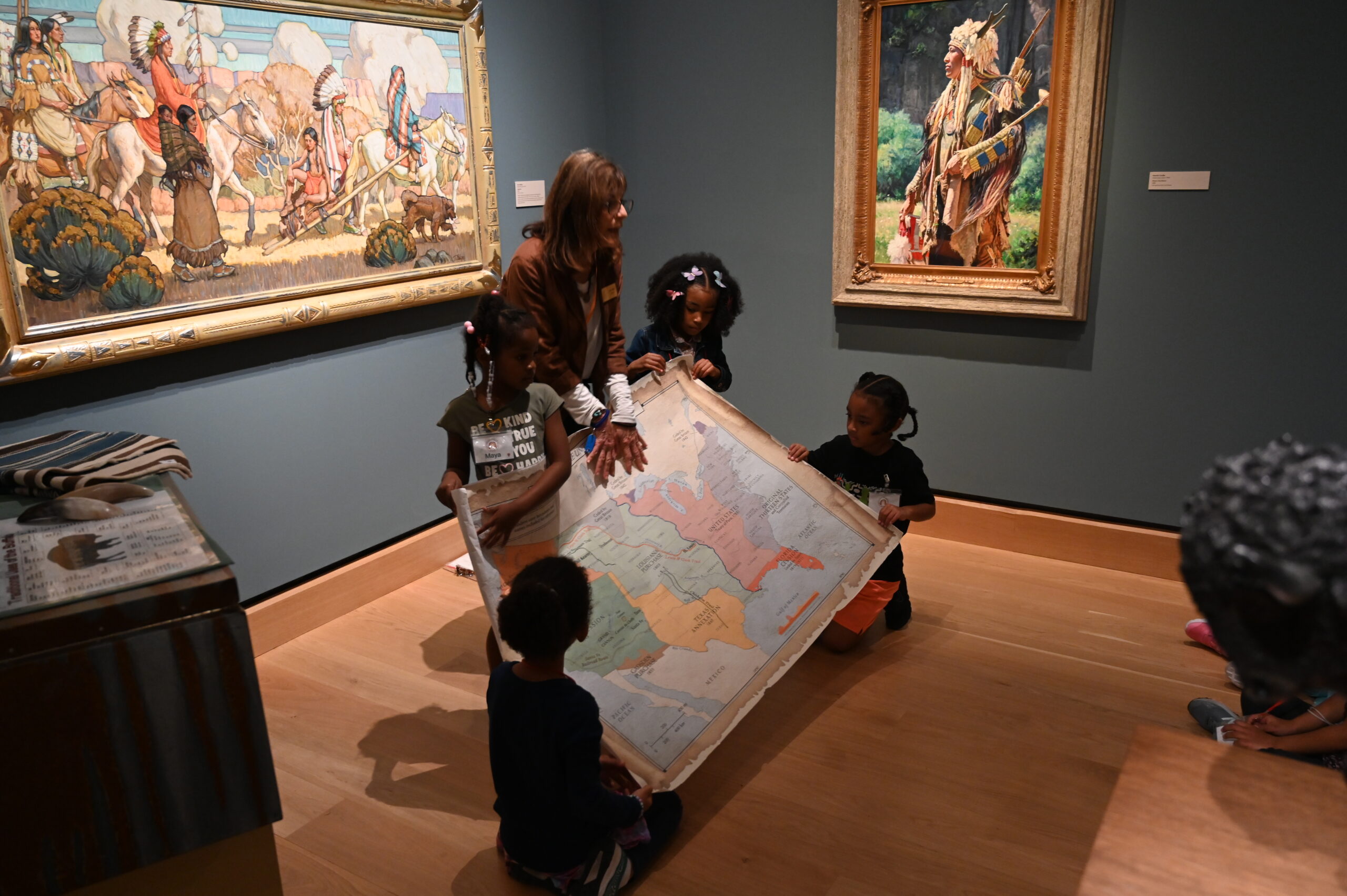 A group of children and an adult examine a large map together during a field trip in an art museum, surrounded by paintings with Native American themes.
