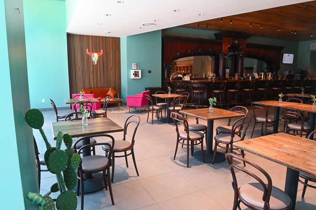 Modern restaurant interior with wooden tables, brown chairs, a bar area, a neon bull skull light, pink armchairs, and a potted cactus—perfect for unique event spaces.