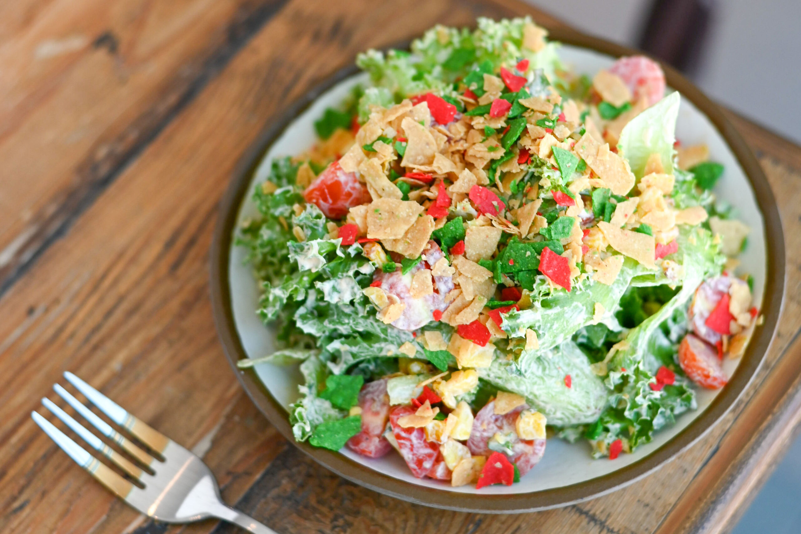A plate of salad with lettuce, tomatoes, herbs, creamy dressing, and crushed tortilla chips on top sits next to a fork on a wooden table—perfect for a quick bite during your lunch hours. Parking is available nearby for your convenience.