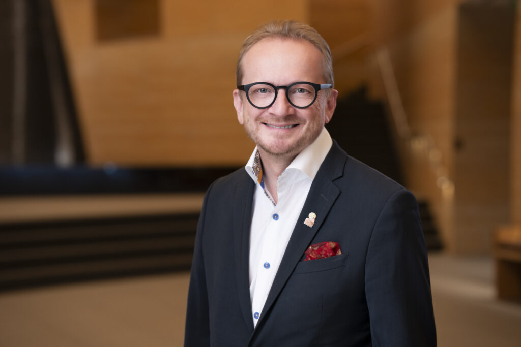A man in a suit and glasses stands indoors, smiling at the camera with a blurred staircase and wooden walls in the background.