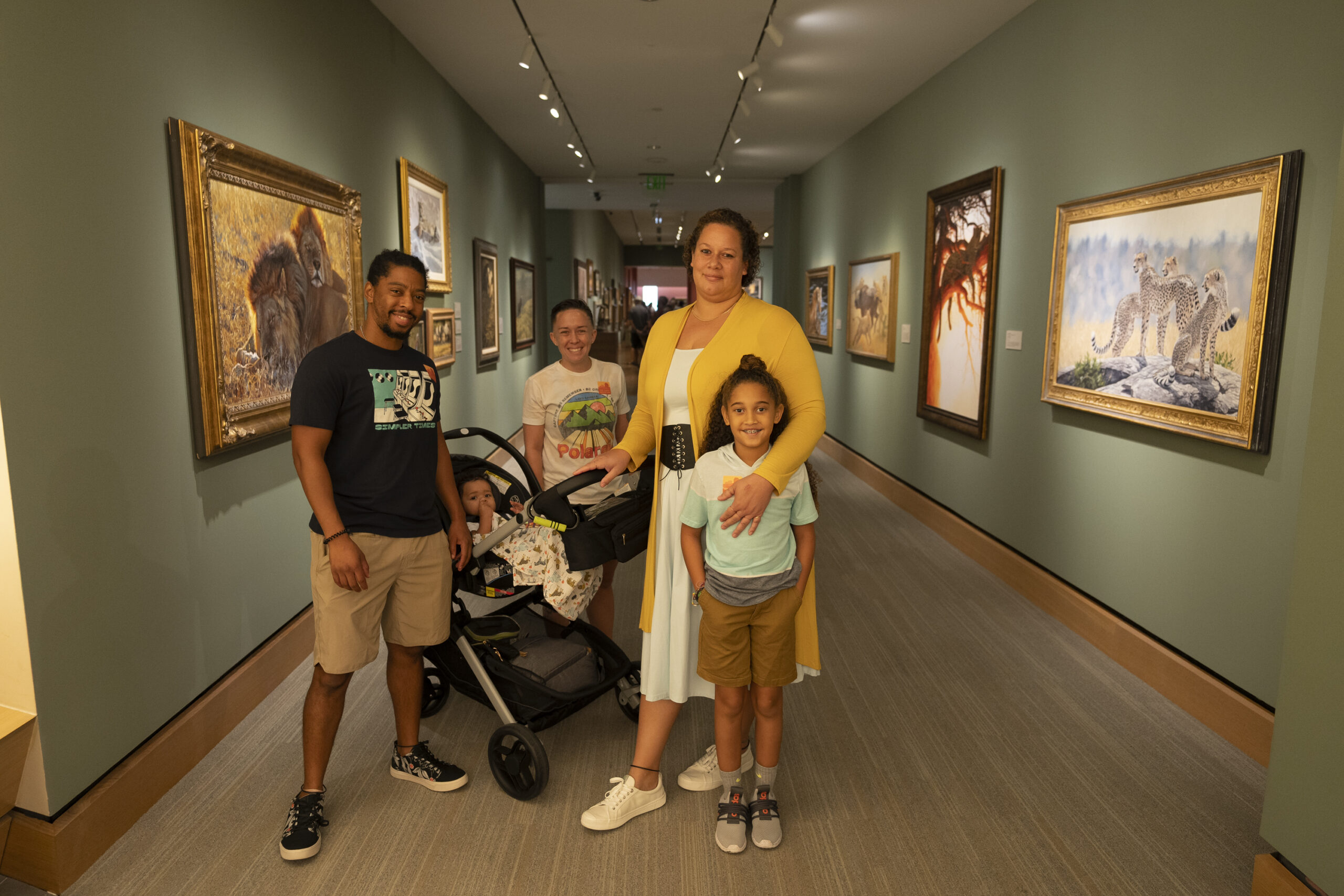 A family of five stands in an art gallery hallway with paintings of animals on the walls. Two children stand with the parents, and one child sits in a stroller.