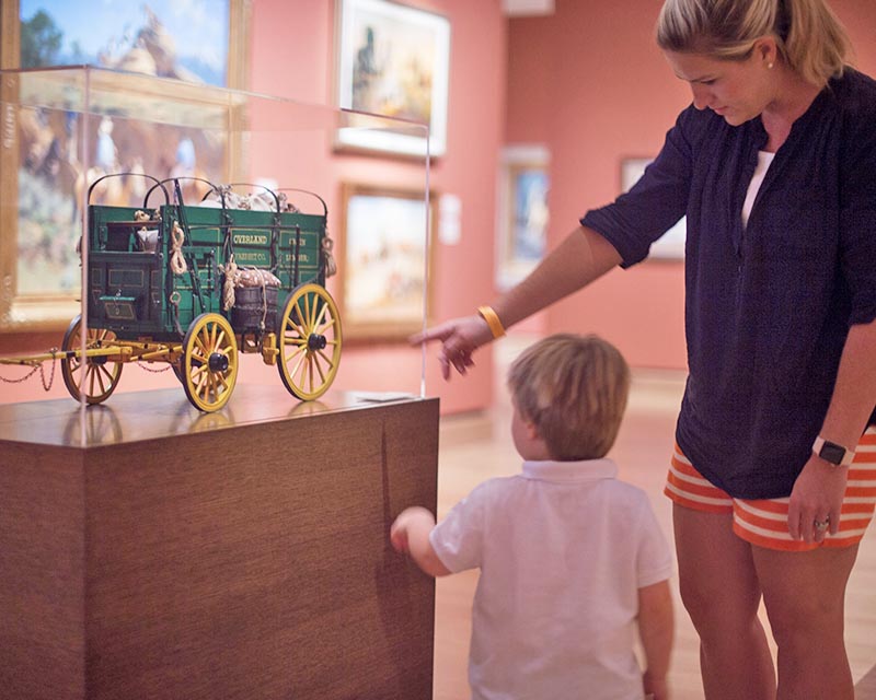 A woman and child view and point at a miniature green wagon displayed in a glass case in an art museum. Support exhibits like this—Make a Donation today.