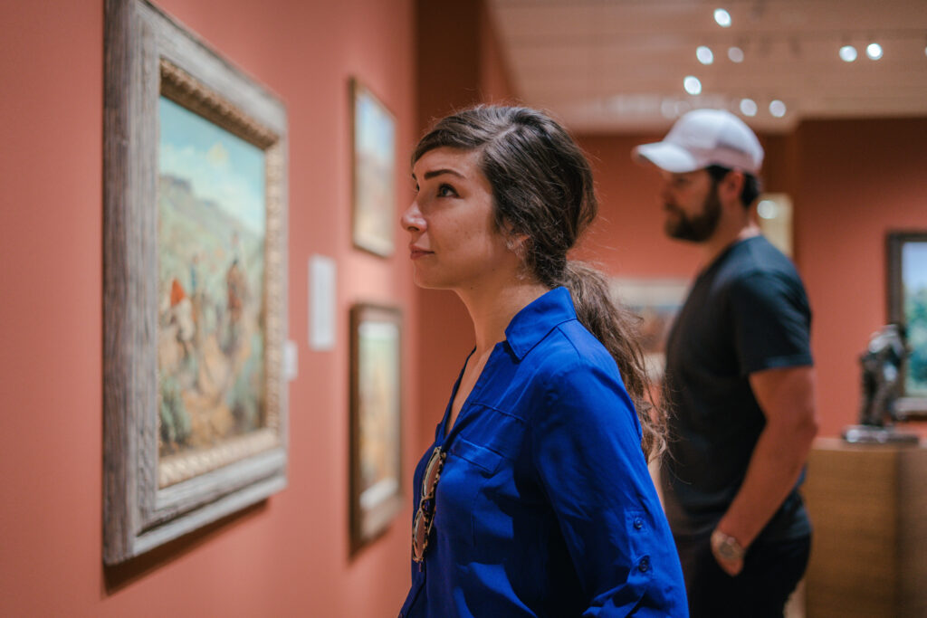 A woman in a blue shirt looks at a painting in an art gallery, while a man in a hat stands in the background.