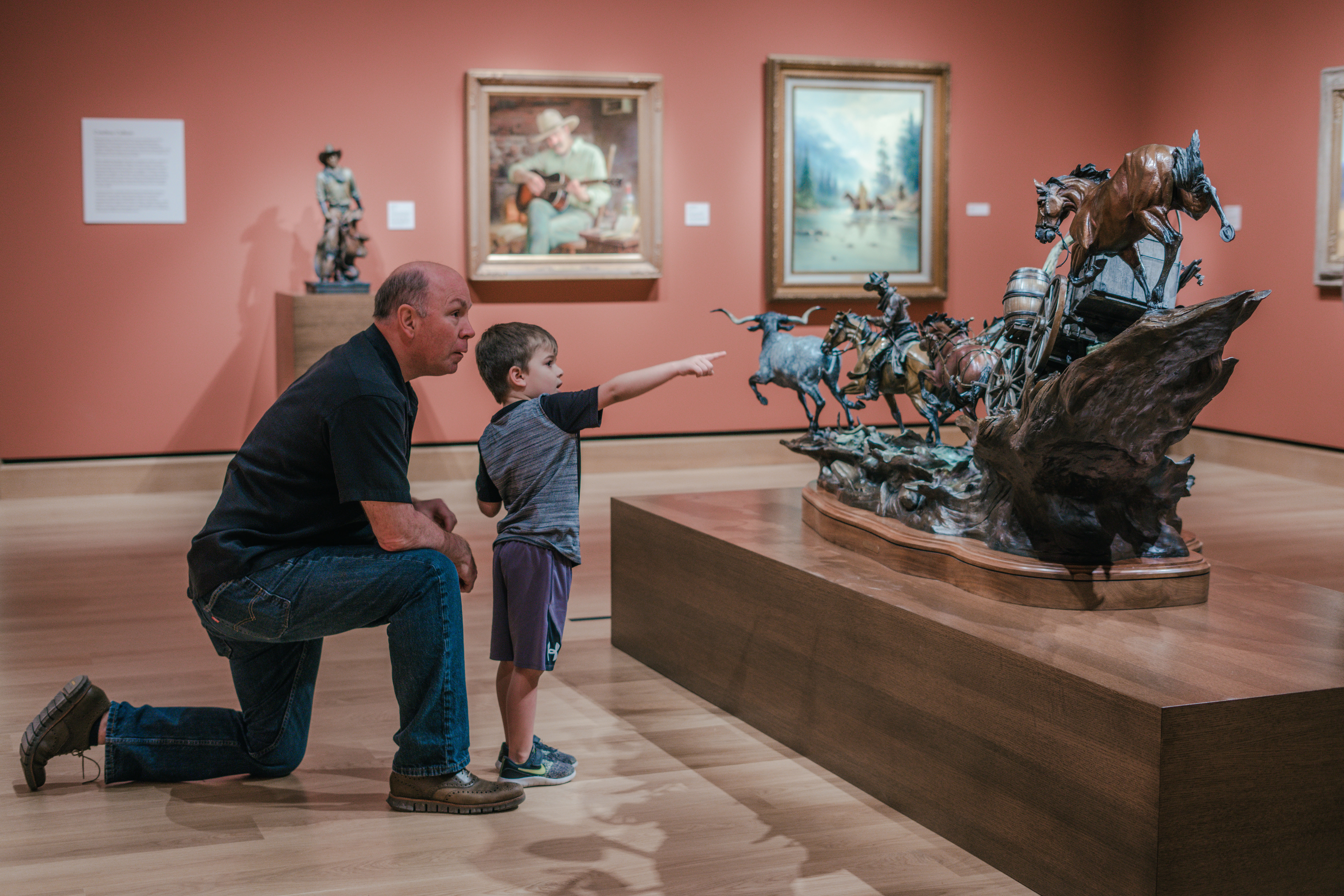 An adult and a child observe a western-themed bronze sculpture in an art museum; the child points at the artwork. Paintings hang on the walls in the background.
