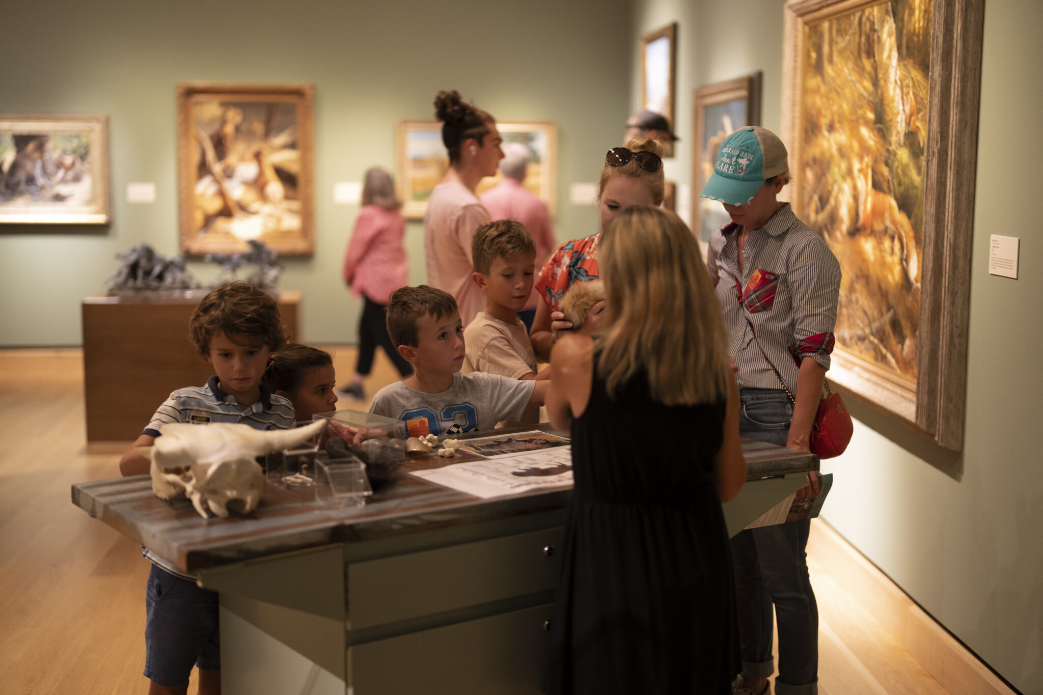 A group of children and adults observe and discuss artifacts on display in an art museum gallery with paintings on the walls.