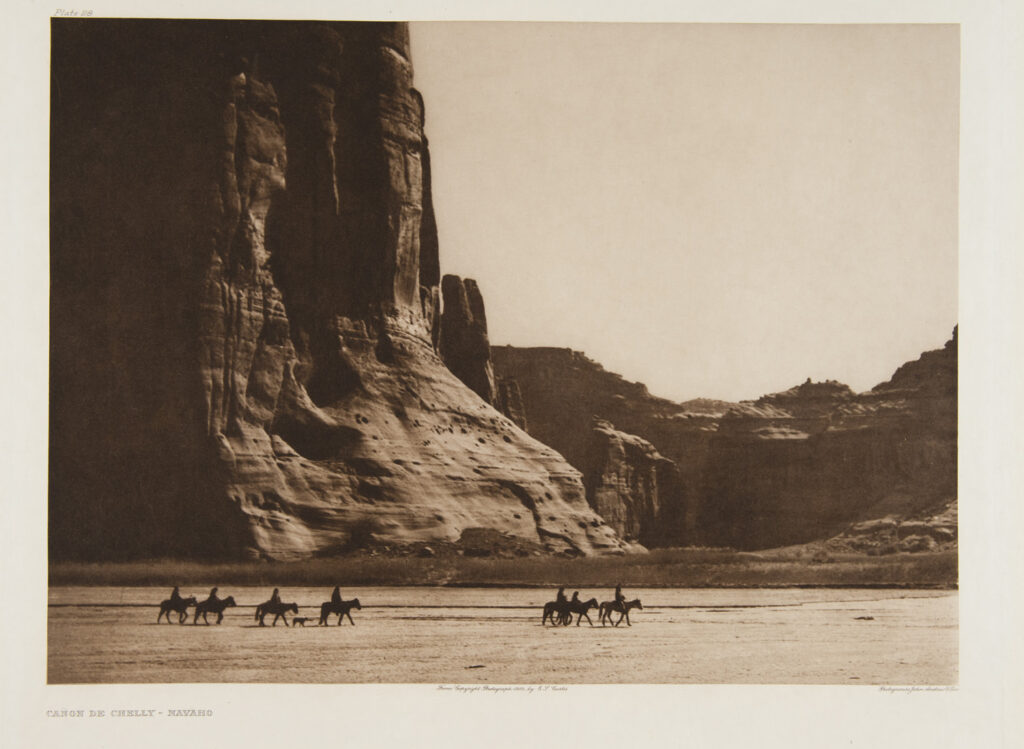 A group of riders on horseback travel across a flat, open plain at the base of large rock cliffs in Canyon de Chelly, Navajo Nation, evoking scenes featured in the Early West Gallery.