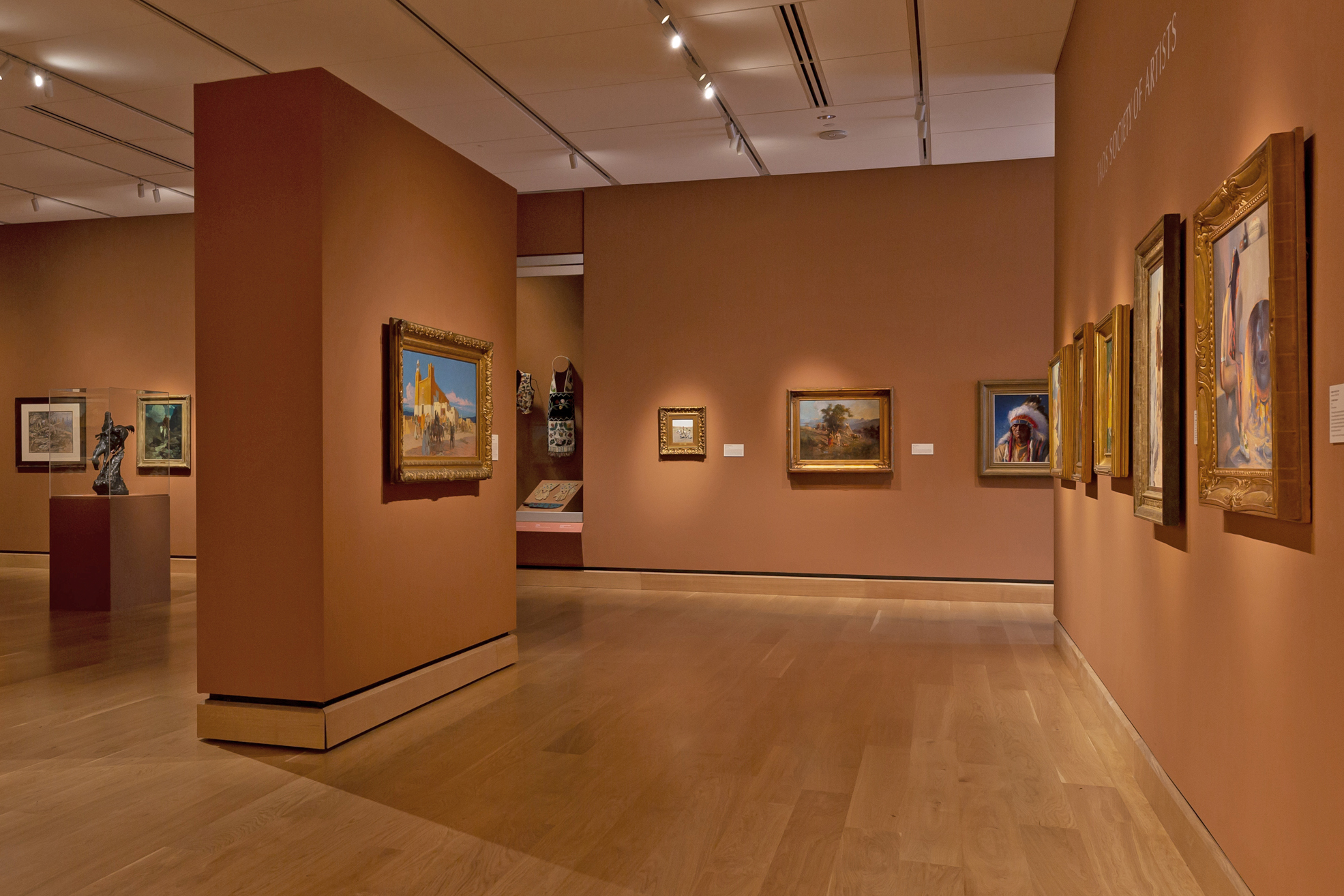Art gallery interior with wooden floors and terracotta walls, displaying framed paintings and sculptures under ceiling lights.