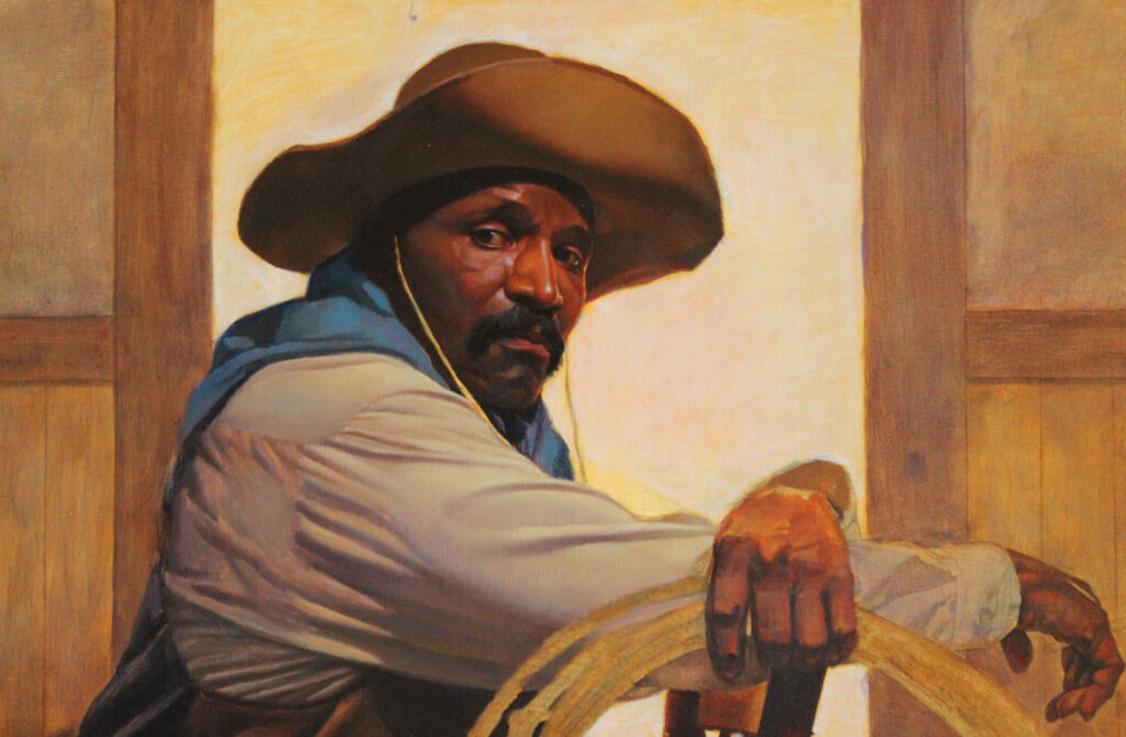 A man wearing a wide-brimmed hat and a blue scarf sits with his arm resting on the back of a wooden chair at the Frontier Gallery, gazing directly forward.