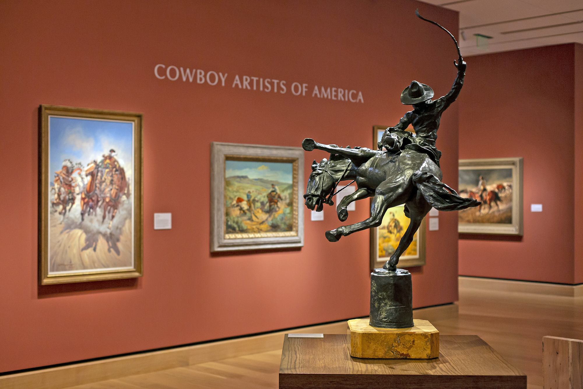 A bronze sculpture of a cowboy on horseback is displayed in front of paintings in a museum gallery labeled "Cowboy Artists of America.
