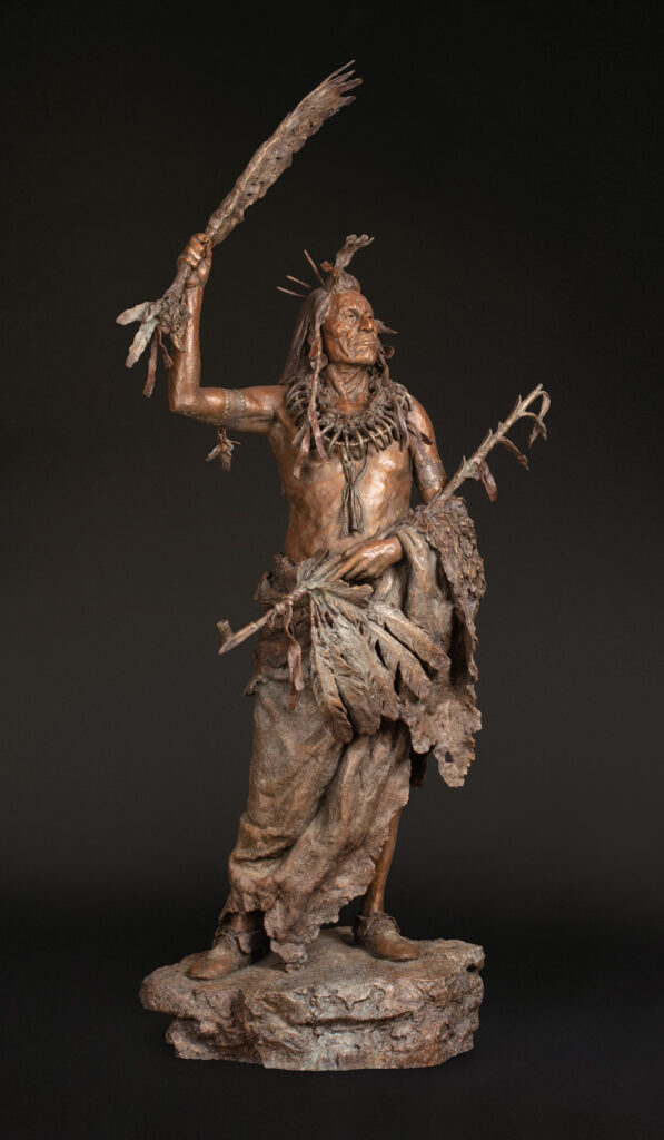 Bronze statue of an Indigenous man in traditional attire holding a feathered staff aloft, with another hand grasping leaves, stands on a textured base in the Introductory Gallery against a dark background.