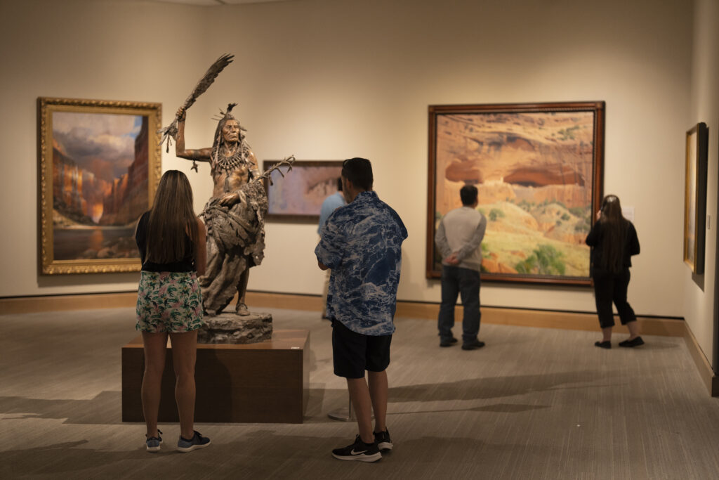 Four people in a museum gallery observe paintings and a bronze Native American sculpture displayed on a pedestal.