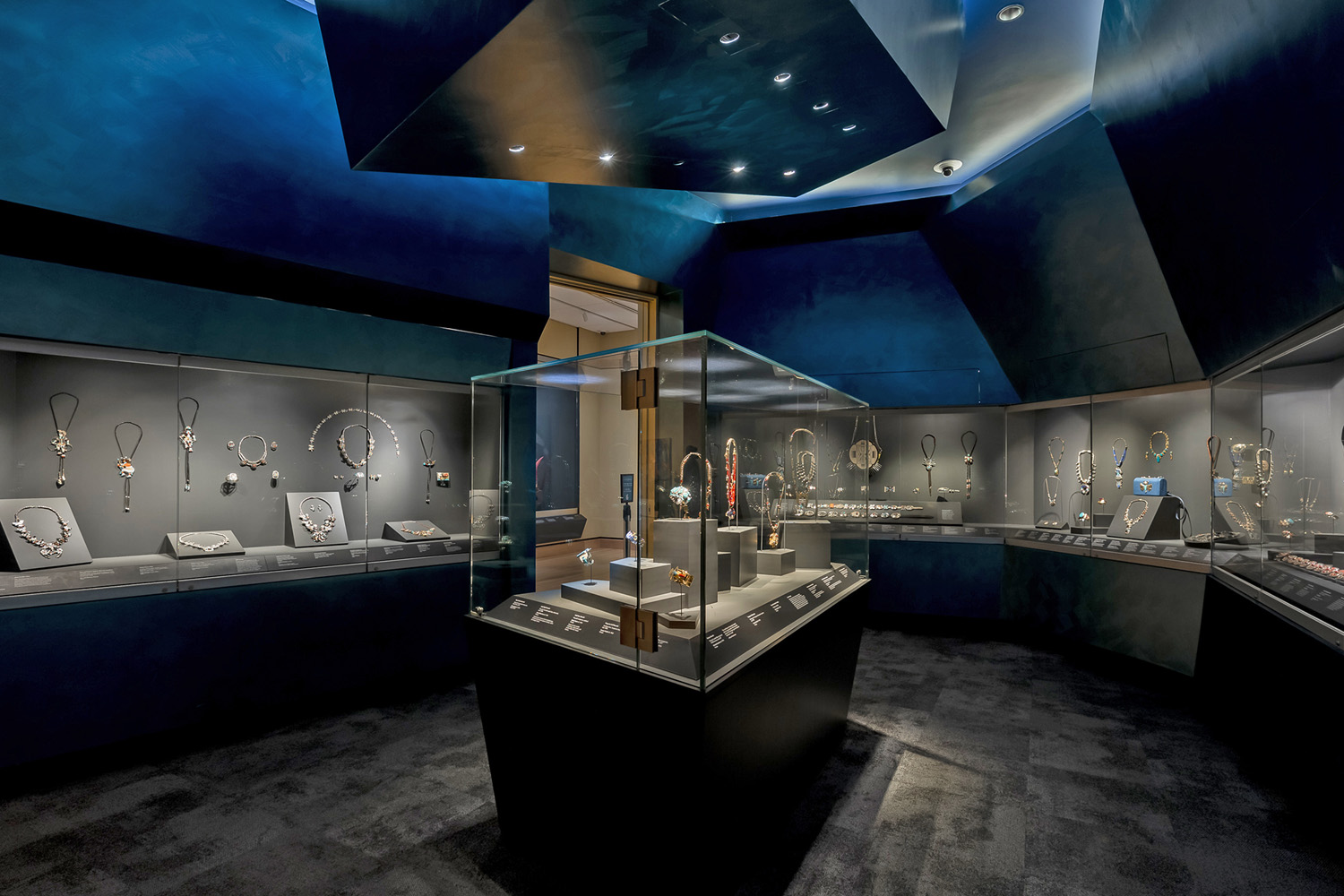 A museum exhibit room resembles a Jewel Box, displaying various pieces of jewelry in glass cases. Necklaces, bracelets, and other adornments are arranged on stands under focused lighting, evoking the elegance of a fine jewelry store.