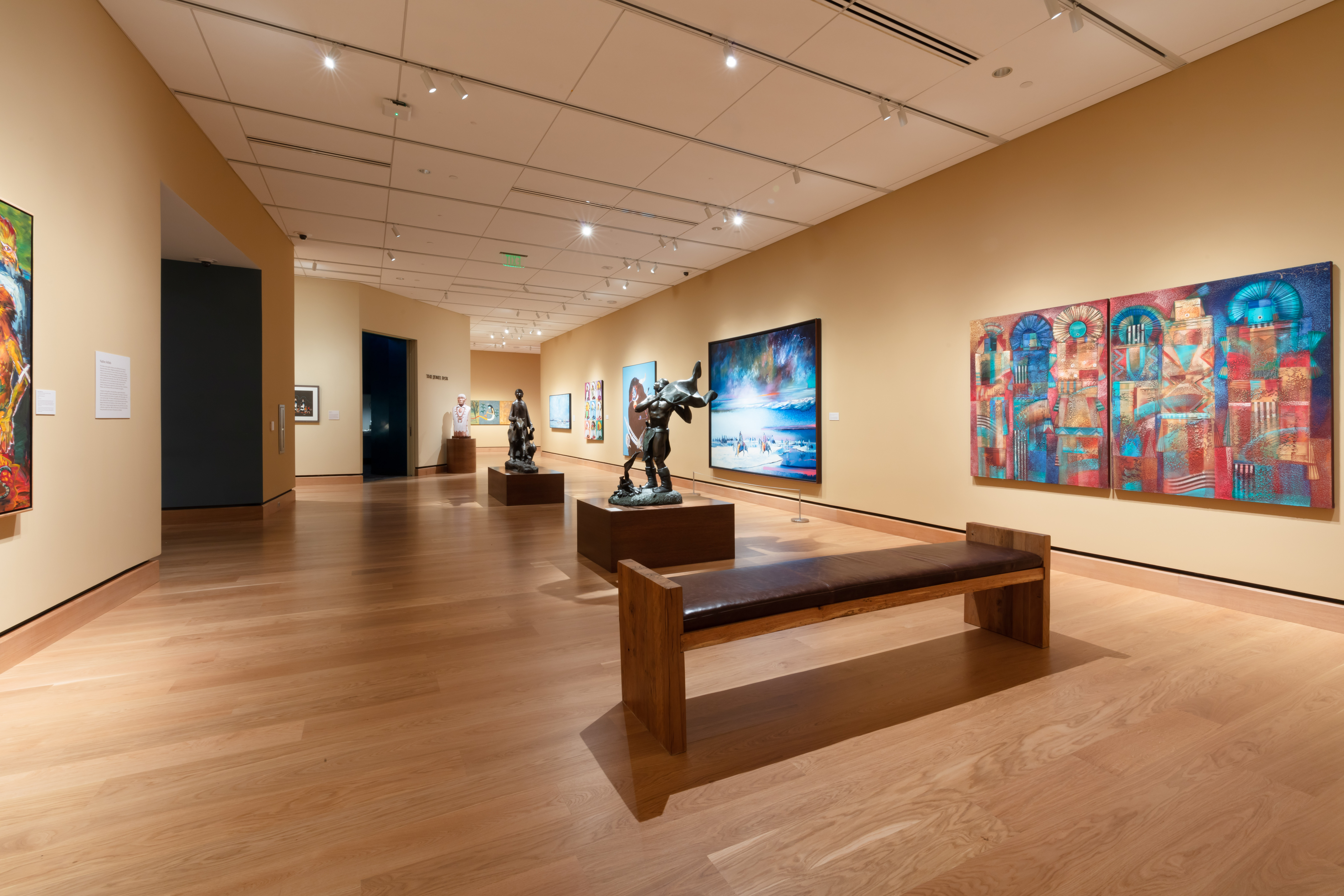Art gallery interior with wooden floors, benches, sculptures, and colorful paintings displayed on beige walls under ceiling spotlights.