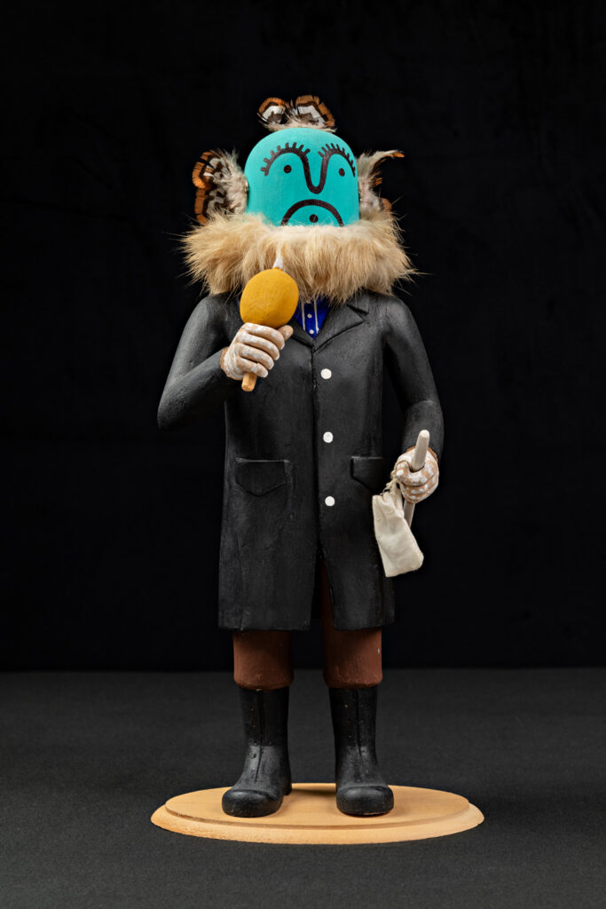 A figurine from Native Life Gallery wears a black coat and fur collar, holds a yellow rattle and white pouch, with a blue mask and butterfly decorations on its head, standing on a round base against a black background.