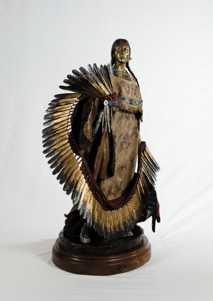 A bronze sculpture of a Native American woman wearing a traditional dress and holding a feathered fan, displayed on a round wooden base, showcased as part of the Native Life Gallery against a white background.