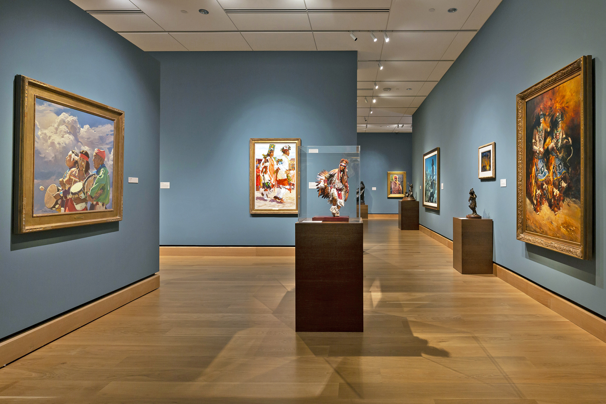Art gallery interior with blue walls, wooden floor, framed paintings on the walls, and sculptures displayed on pedestals.