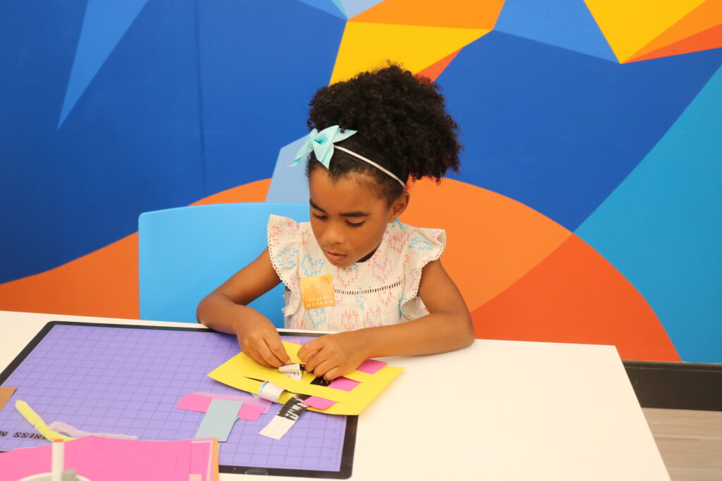 A young girl sits at a table making a craft project with colorful paper, in front of a blue geometric-patterned wall.