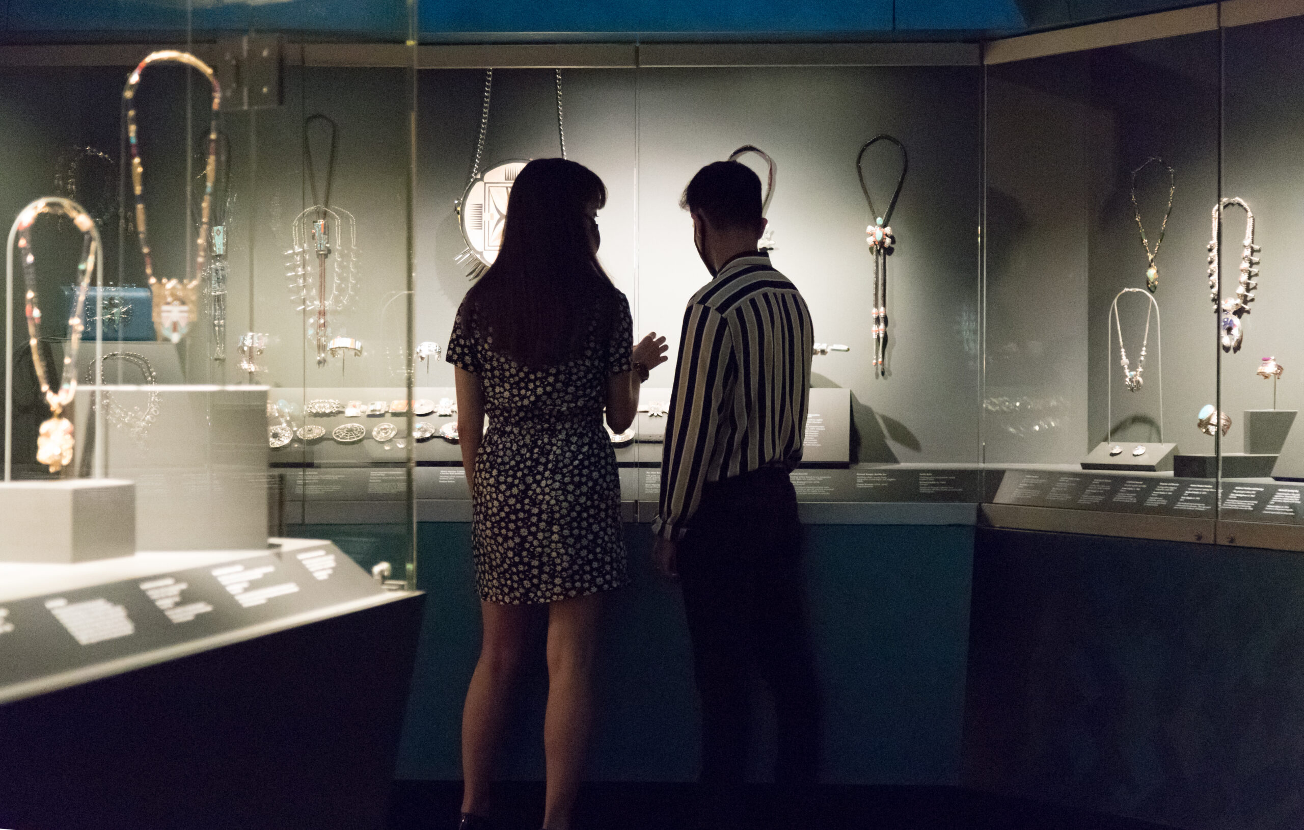 Two people stand in front of a glass display case observing various necklaces and jewelry pieces in a dimly lit museum or exhibition setting.