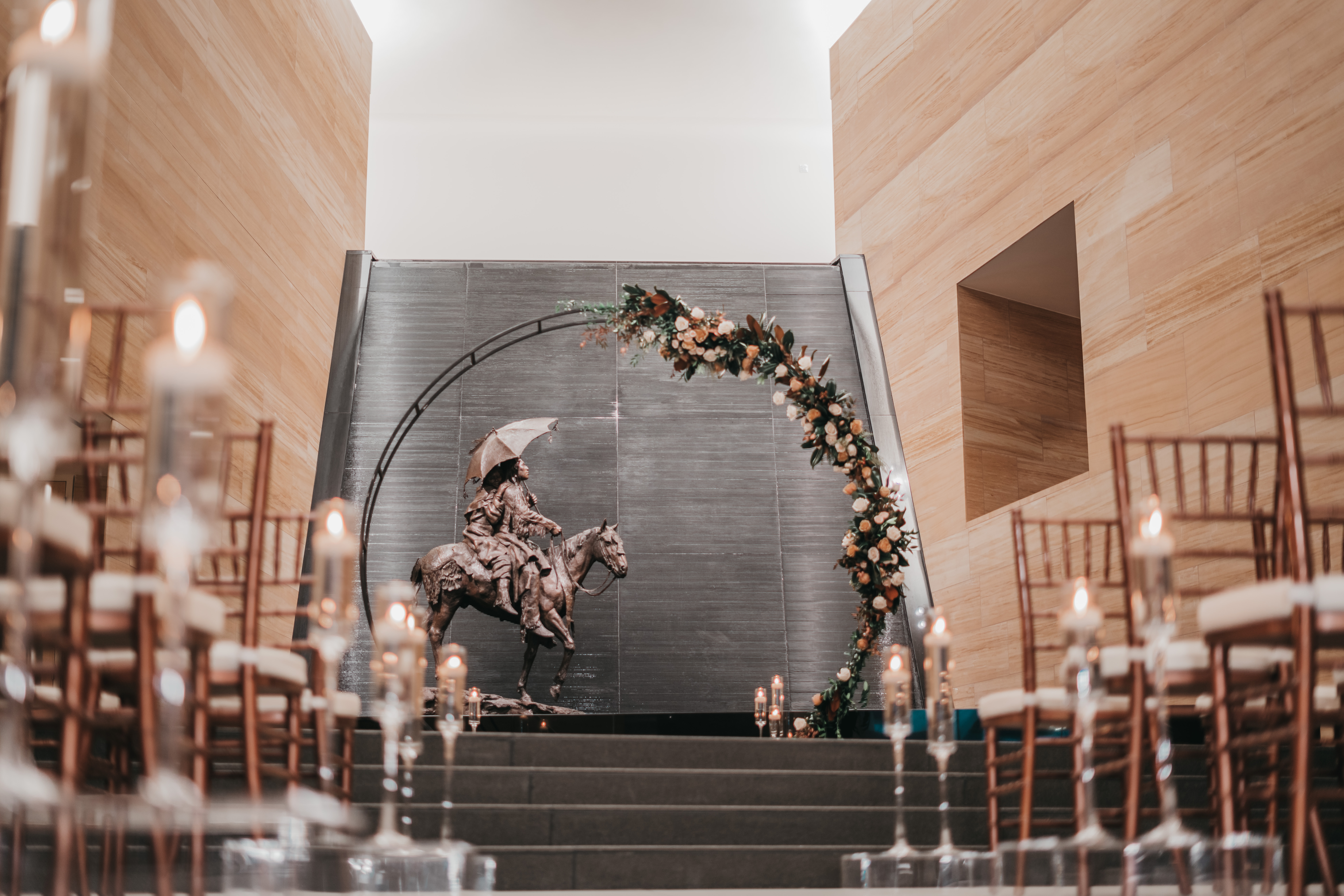 A bronze statue of a rider on horseback stands behind a floral circular arch at the end of an aisle lined with chairs and candles in one of the city's most unique event spaces, set within a modern indoor setting.