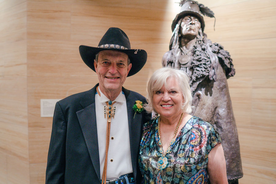 An older man in a suit and cowboy hat stands beside an older woman in a colorful dress, with a bronze Native American statue in the background.