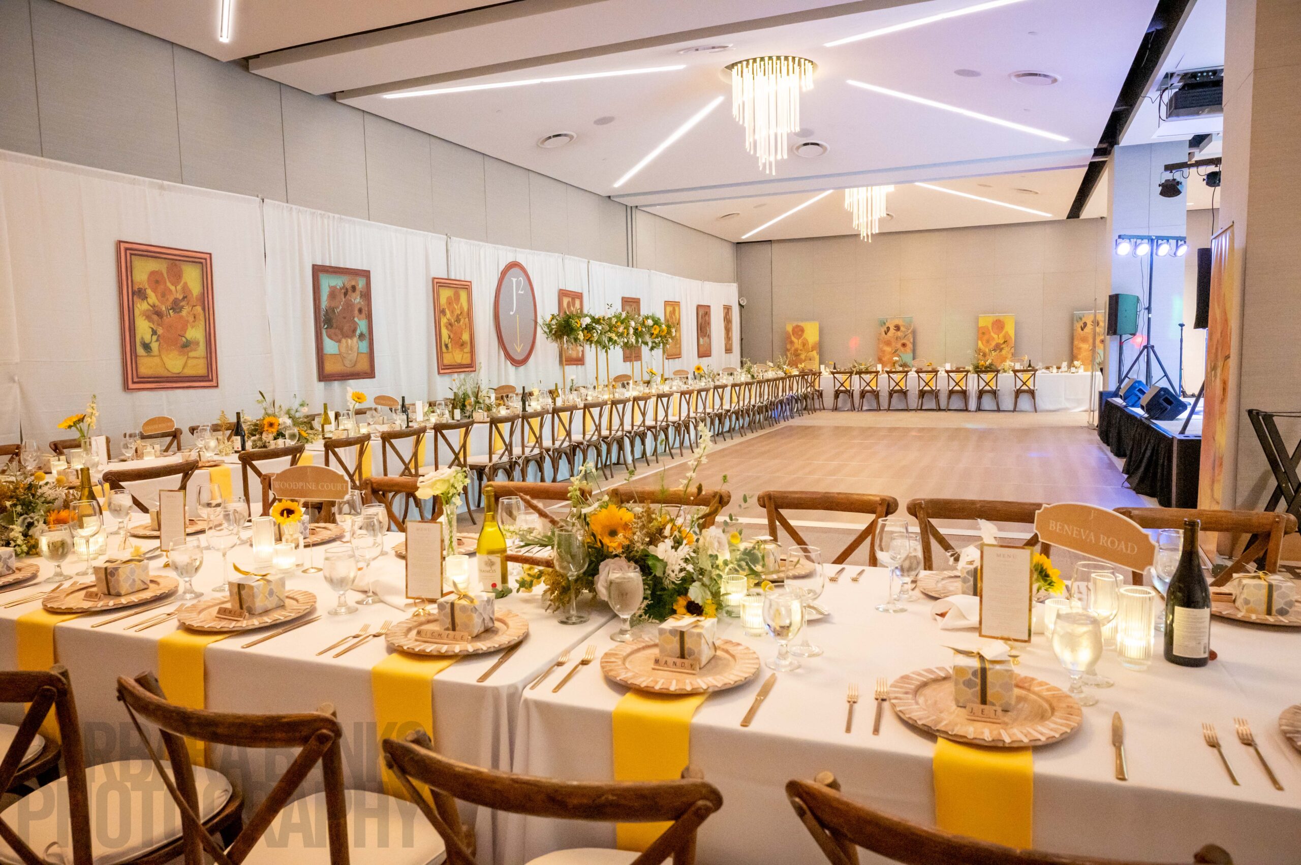 A banquet hall is set with long tables, yellow-themed decor, sunflowers, place settings, and glassware, ideal for meetings or corporate events, with framed artwork on the walls and a chandelier overhead.