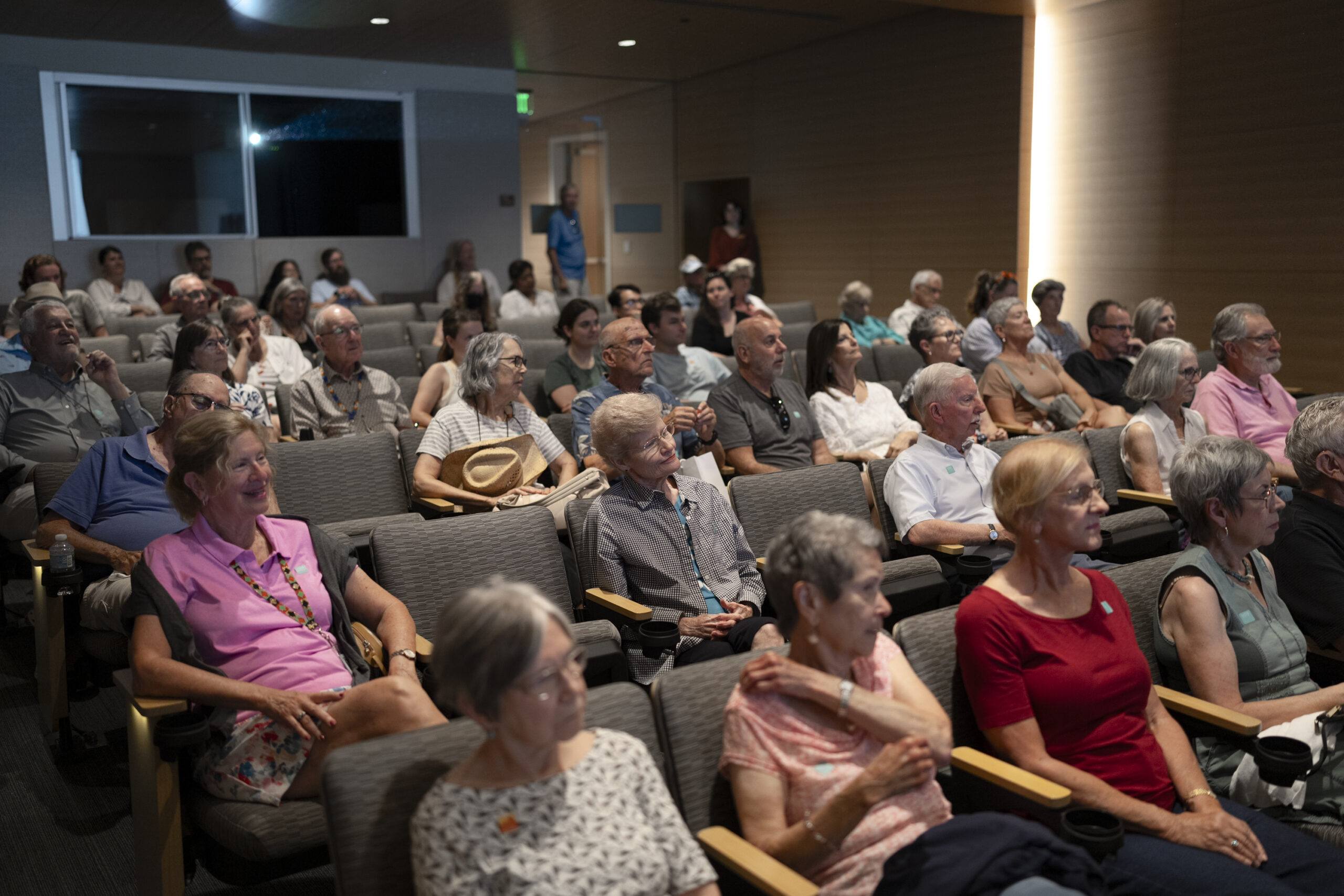 A group of older adults sit in rows of chairs, attentively watching a presentation or event in a dimly lit auditorium, showcasing the versatility of event spaces designed for engaging community gatherings.