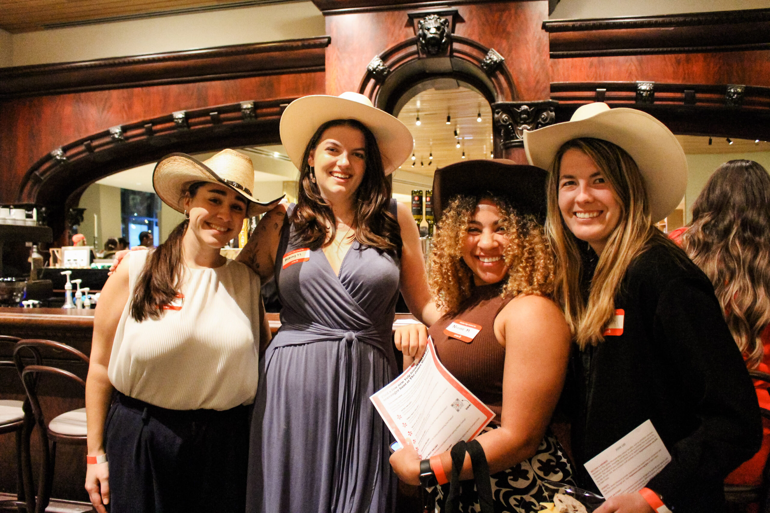 Four women wearing cowboy hats stand together and smile indoors, each holding papers and wearing name tags, with a wooden bar in the background.