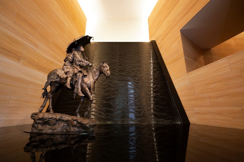 Bronze statue of a rider on horseback holding an umbrella, positioned in front of a modern indoor water feature with wooden paneled walls.
