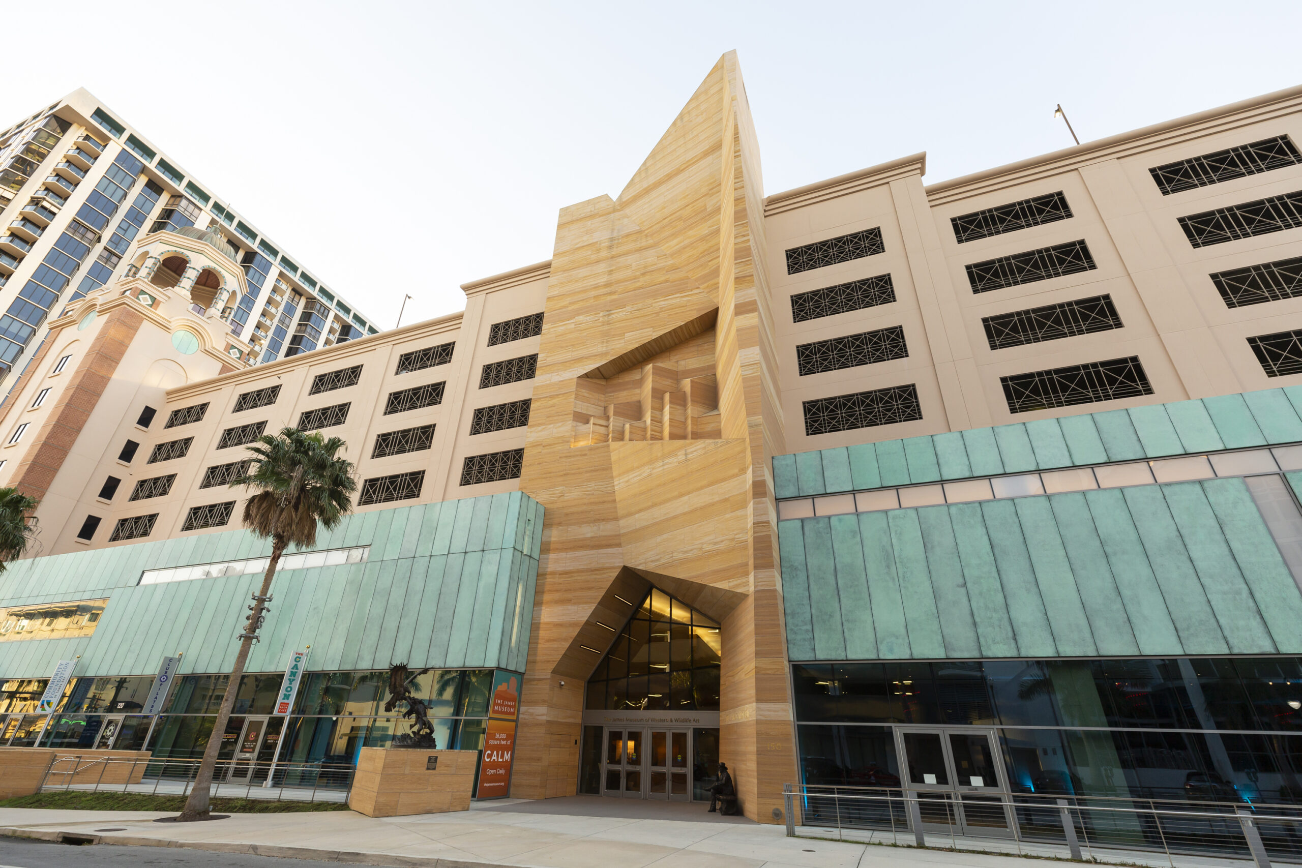 Modern building with tan stone geometric entrance, teal-green panels, glass doors, and palm trees in front; multi-story structure with decorative metal grates and high-rise nearby.