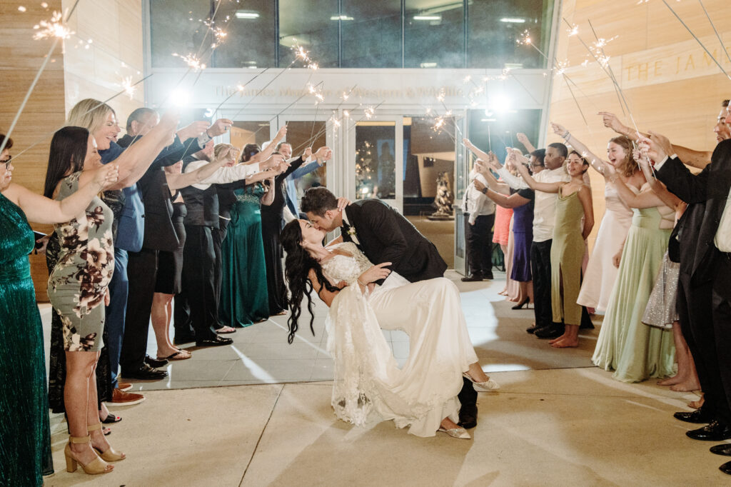 A bride and groom share a dip kiss outside stunning event spaces as guests on both sides hold sparklers, celebrating at night in formal attire.