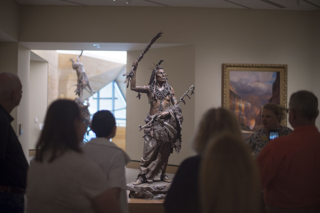 A group of people observes a bronze statue of a Native American man holding a sword and staff in a museum gallery.