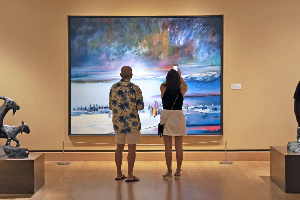 Two people stand in front of a large, colorful landscape painting in an art gallery; one person takes a photo with a smartphone.